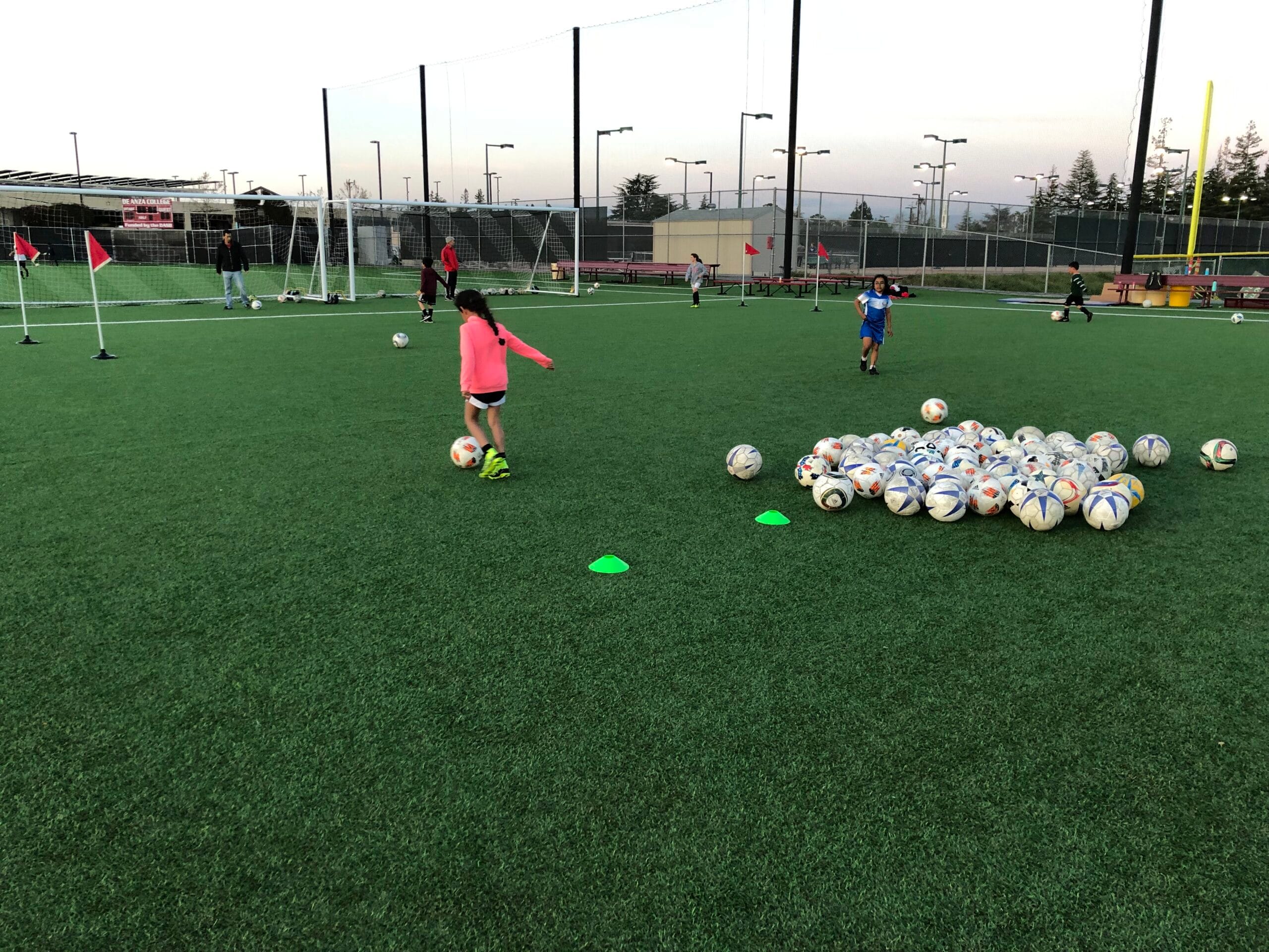 Young athletes participating in technical ball control drills and finishing practice during a Cupertino FC camp on a turf field. There are dozens of balls next to them.