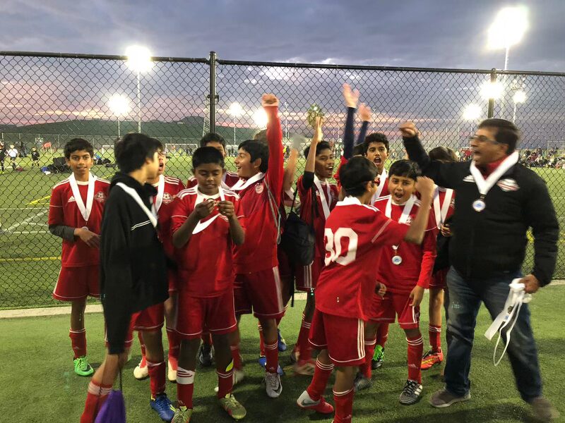 A Cupertino FC youth soccer team and their coach celebrating with medals under stadium lights after a successful tournament win.